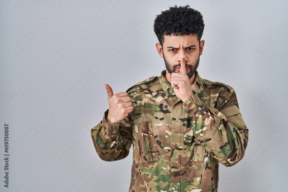 Arab man wearing camouflage army uniform asking to be quiet with finger on lips pointing with hand to the side. silence and secret concept.