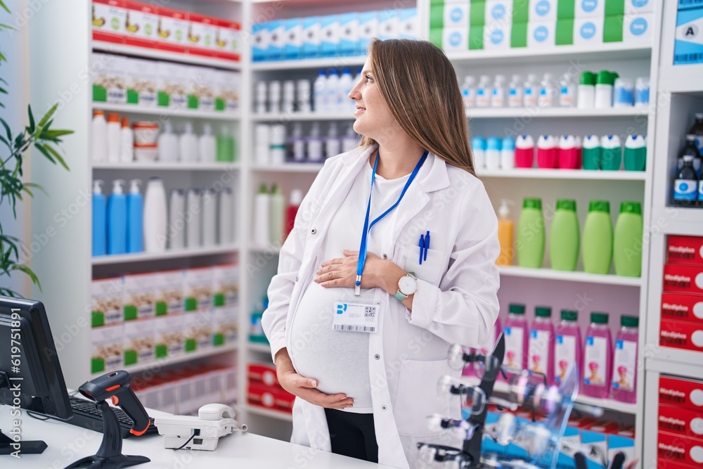 Young pregnant woman pharmacist smiling confident touching belly at pharmacy