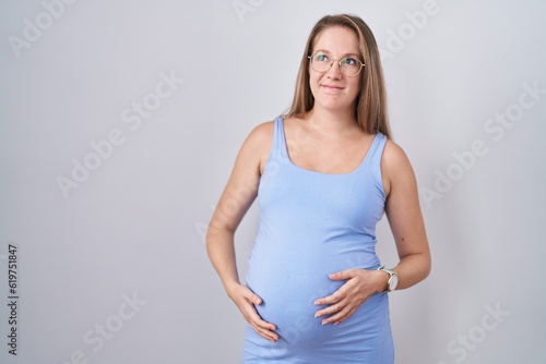 Young pregnant woman standing over white background smiling looking to the side and staring away thinking.