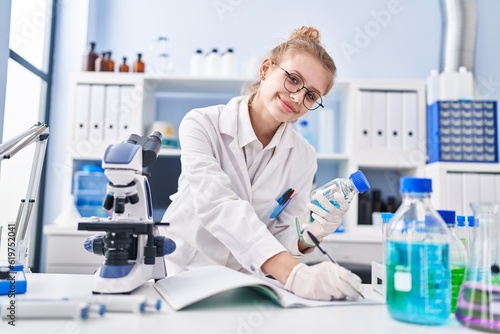 Young blonde woman scientist writing on notebook holding bottle at laboratory
