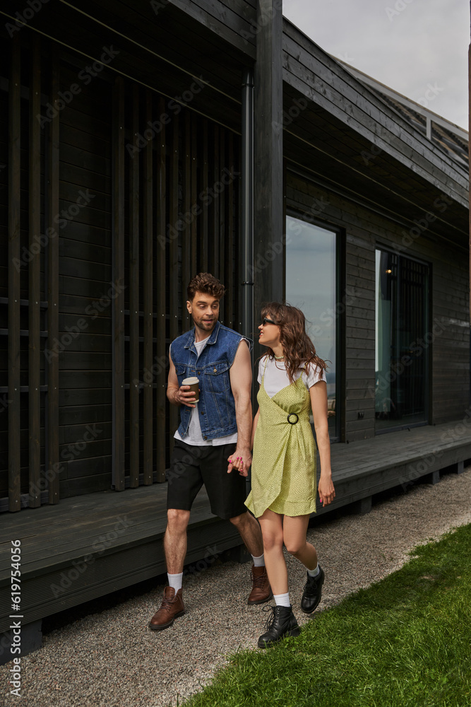 Fototapeta premium Stylish man in summer outfit holding coffee to go and hand of cheerful girlfriend in sunglasses and sundress while walking near wooden house in rural setting, outdoor enjoyment concept, tranquility