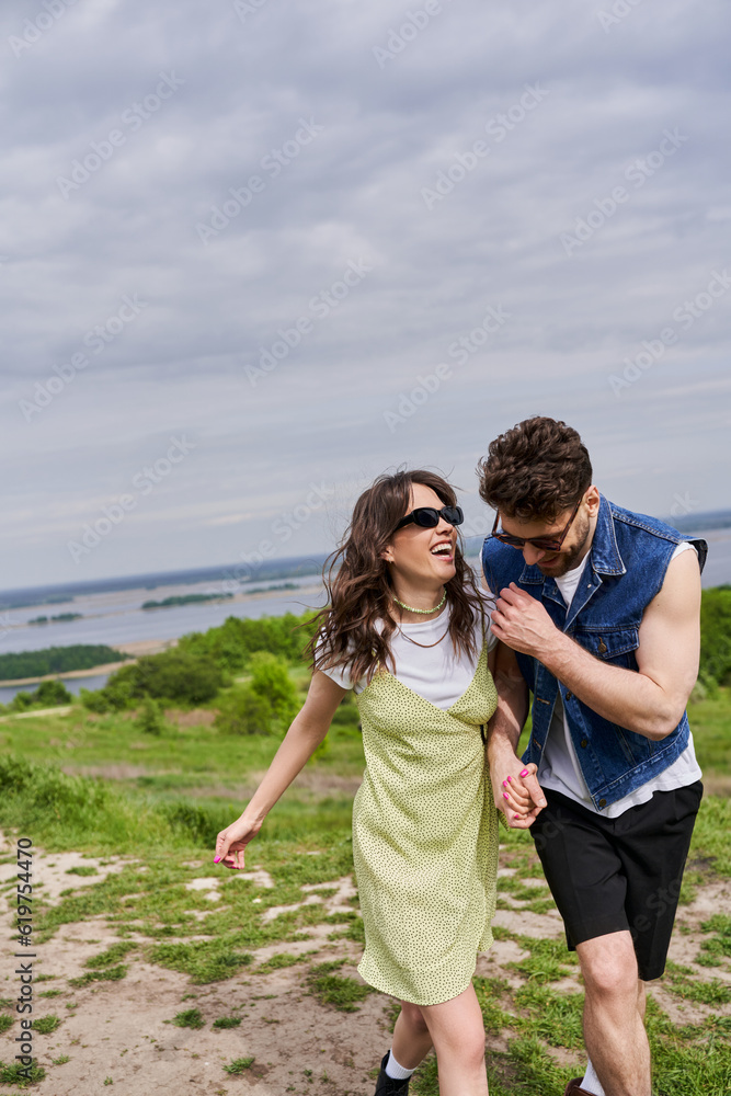 Cheerful brunette man in stylish summer outfit and denim vest holding hand of excited girlfriend in sunglasses and sundress and walking together on path on grassy hill, countryside leisurely stroll