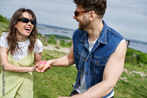 Cheerful brunette woman in trendy summer outfit and sunglasses holding hand of boyfriend in denim vest while spending time in blurred rural setting at background, countryside leisurely stroll © LIGHTFIELD STUDIOS