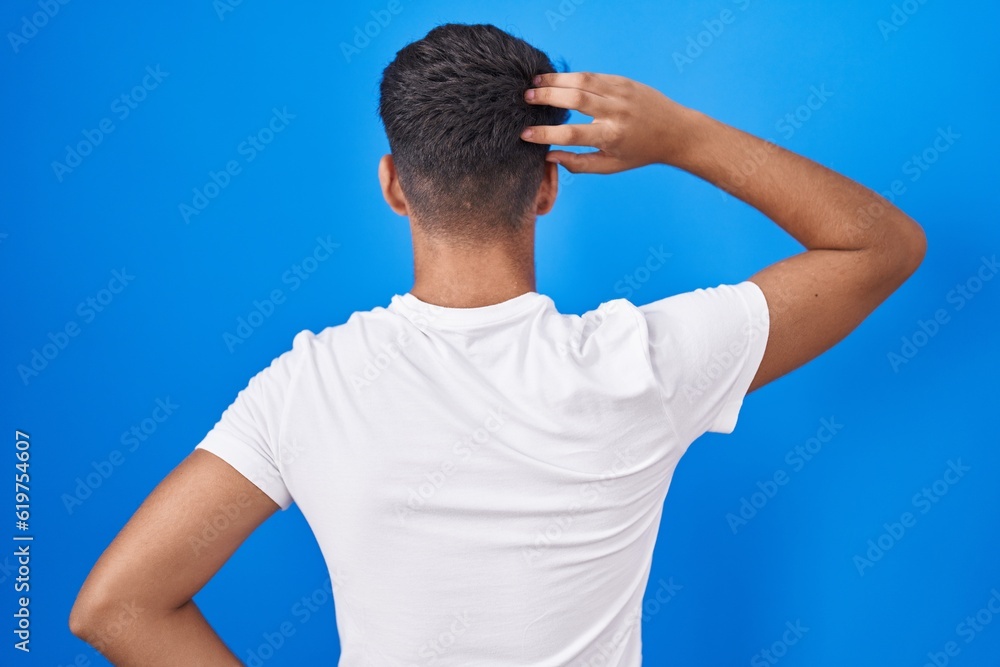 Hispanic teenager standing over blue background backwards thinking about doubt with hand on head Hispanic teenager standing over blue background backwards thinking about doubt with hand on head
