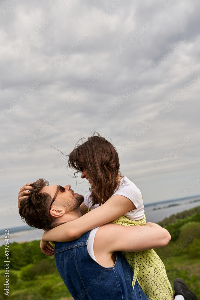 Side view of smiling and trendy man in sunglasses and denim vest hugging and lifting brunette girlfriend in sundress while spending time in rural setting, love story and countryside adventure Side view of smiling and trendy man in sunglasses and denim vest hugging and lifting brunette girlfriend in sundress while spending time in rural setting, love story and countryside adventure