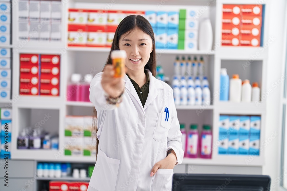 Chinese woman pharmacist smiling confident holding pills bottle at pharmacy