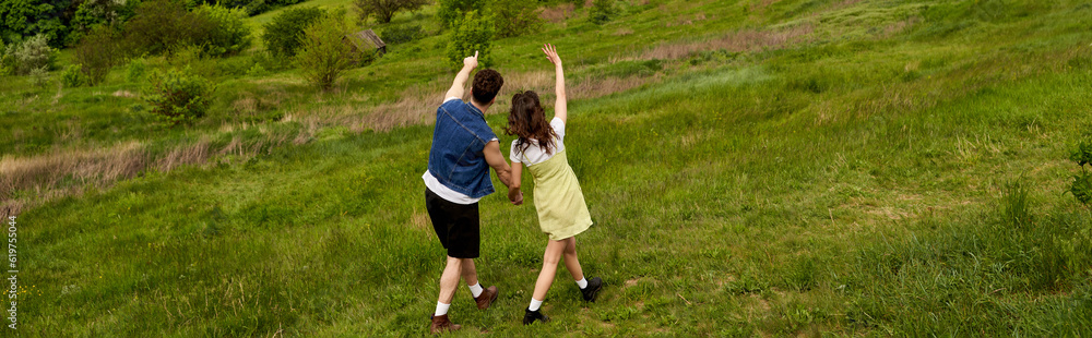 Obraz premium Back view of man in denim vest pointing with finger while holding hand of brunette girlfriend in sundress and standing together on hill with green grass, countryside adventure and love story, banner