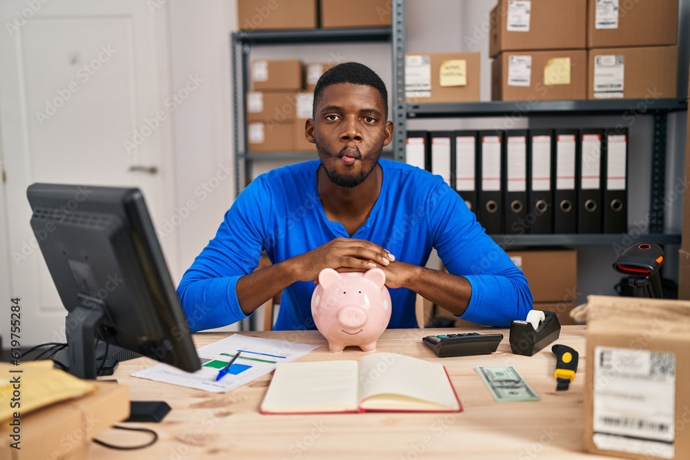 African american man working at small business ecommerce with piggy bank making fish face with mouth and squinting eyes, crazy and comical.