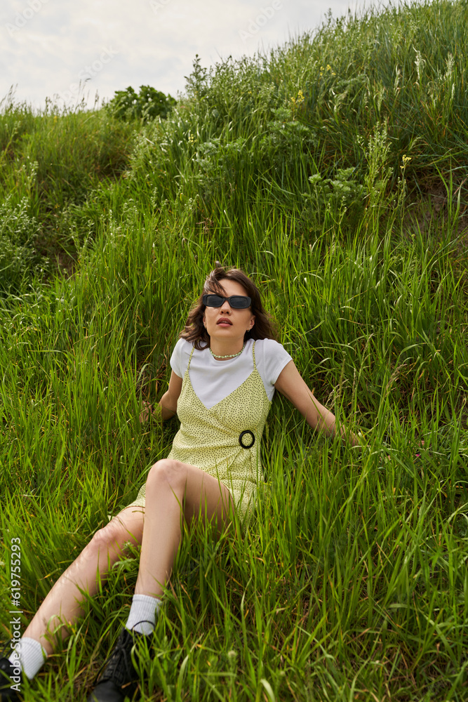 Fashionable brunette woman in sunglasses and sundress spending time and relaxing on green meadow with grass, peaceful retreat and relaxing in nature concept, stylish woman Fashionable brunette woman in sunglasses and sundress spending time and relaxing on green meadow with grass, peaceful retreat and relaxing in nature concept, stylish woman