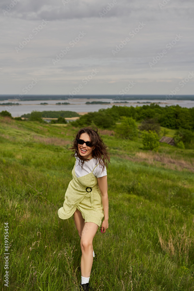 Cheerful and stylish brunette woman in sunglasses and sundress standing on green meadow with grass and spending time with blurred landscape and sky at background, summertime joy Cheerful and stylish brunette woman in sunglasses and sundress standing on green meadow with grass and spending time with blurred landscape and sky at background, summertime joy