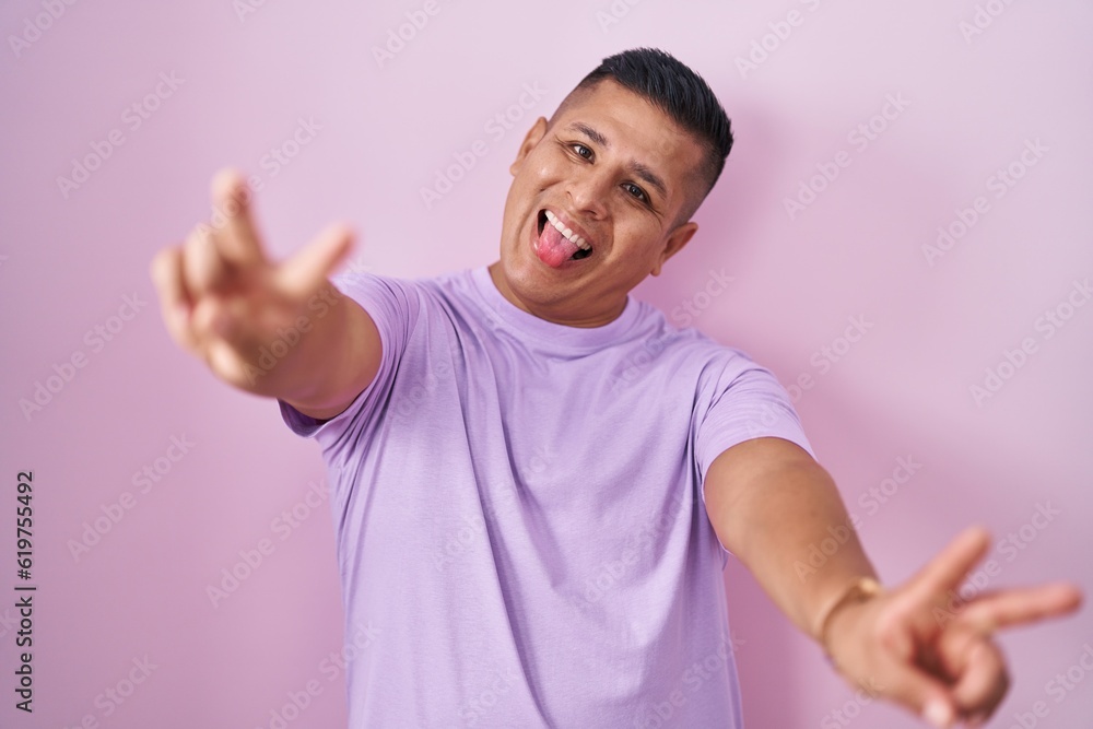 Young hispanic man standing over pink background smiling with tongue out showing fingers of both hands doing victory sign. number two. Young hispanic man standing over pink background smiling with tongue out showing fingers of both hands doing victory sign. number two.