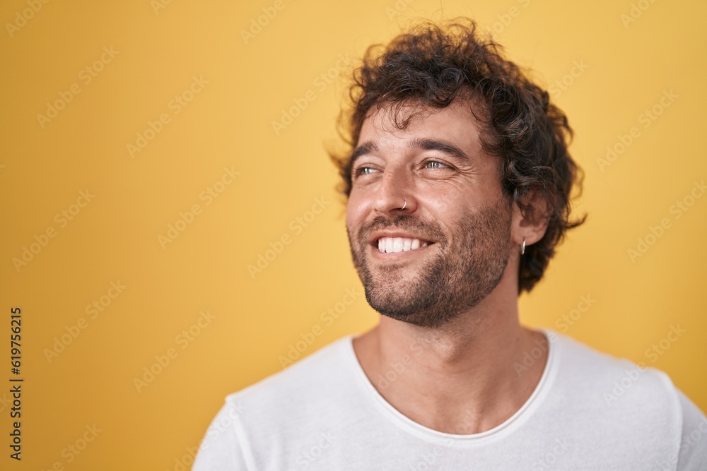 Young hispanic man smiling confident looking to the side over isolated yellow background Young hispanic man smiling confident looking to the side over isolated yellow background