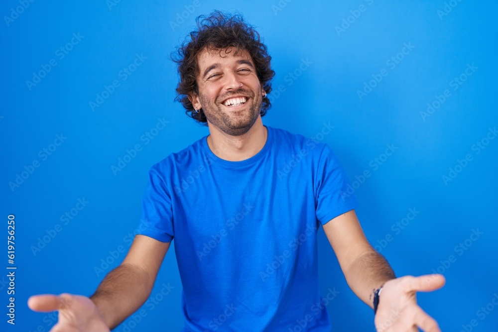 Hispanic young man standing over blue background smiling cheerful with open arms as friendly welcome, positive and confident greetings Hispanic young man standing over blue background smiling cheerful with open arms as friendly welcome, positive and confident greetings