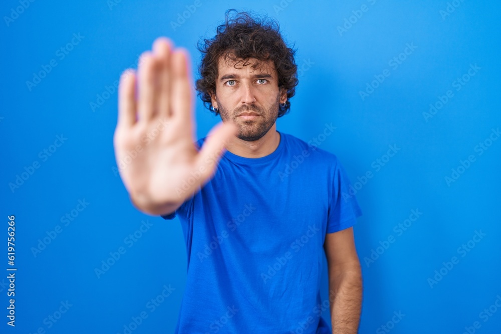 Hispanic young man standing over blue background doing stop sing with palm of the hand. warning expression with negative and serious gesture on the face. Hispanic young man standing over blue background doing stop sing with palm of the hand. warning expression with negative and serious gesture on the face.