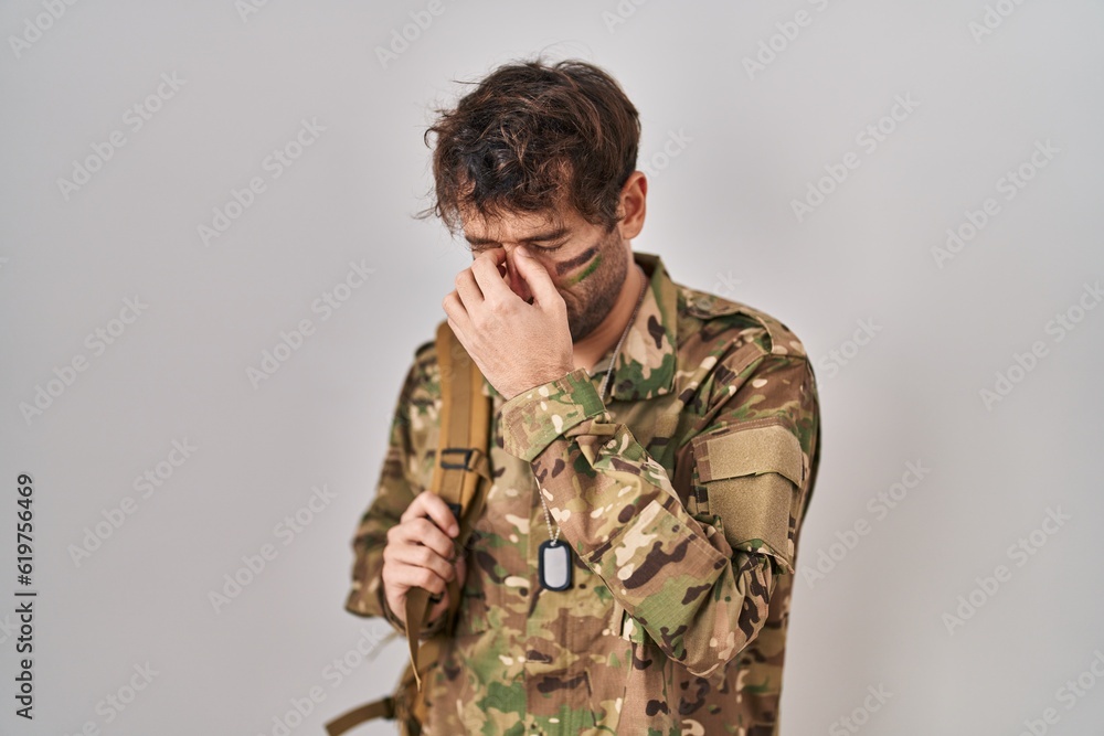 Hispanic young man wearing camouflage army uniform tired rubbing nose and eyes feeling fatigue and headache. stress and frustration concept.