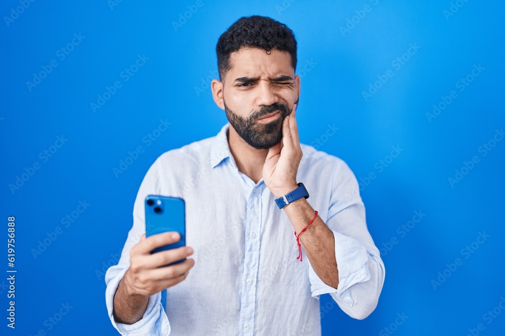 Hispanic man with beard using smartphone typing message touching mouth with hand with painful expression because of toothache or dental illness on teeth. dentist concept. Hispanic man with beard using smartphone typing message touching mouth with hand with painful expression because of toothache or dental illness on teeth. dentist concept.