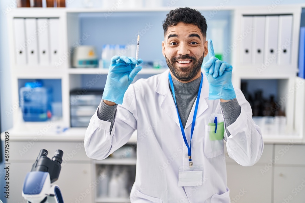 Hispanic man with beard working at scientist laboratory holding syringe smiling with an idea or question pointing finger with happy face, number one Hispanic man with beard working at scientist laboratory holding syringe smiling with an idea or question pointing finger with happy face, number one