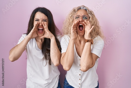 Mother and daughter standing together over pink background shouting angry out loud with hands over mouth