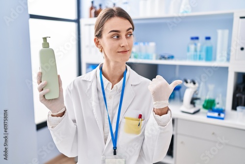 Young caucasian woman working at scientist laboratory holding body lotion pointing thumb up to the side smiling happy with open mouth