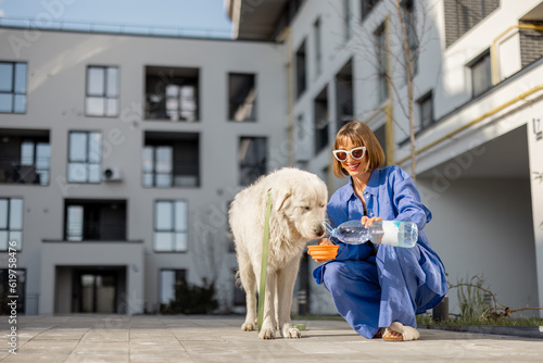 Young woman gives water to drink into portable waterer for her dog during a walk at inner yard of apartment building. concept of pet care