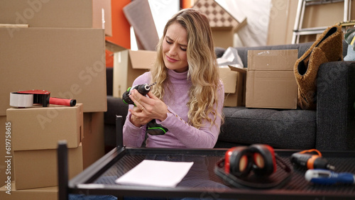 Young blonde woman repairing table using drill with worried expression at new home
