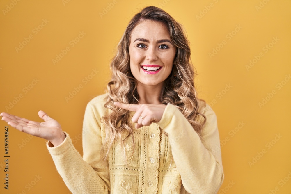Young caucasian woman standing over yellow background amazed and smiling to the camera while presenting with hand and pointing with finger. Young caucasian woman standing over yellow background amazed and smiling to the camera while presenting with hand and pointing with finger.