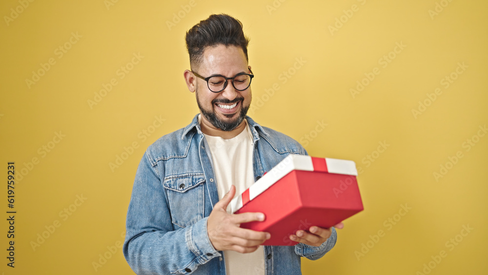 Young latin man smiling confident holding gift over isolated yellow background Young latin man smiling confident holding gift over isolated yellow background