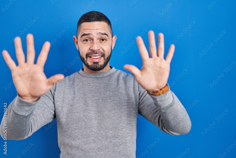 Hispanic man standing over blue background showing and pointing up with fingers number ten while smiling confident and happy. Hispanic man standing over blue background showing and pointing up with fingers number ten while smiling confident and happy.