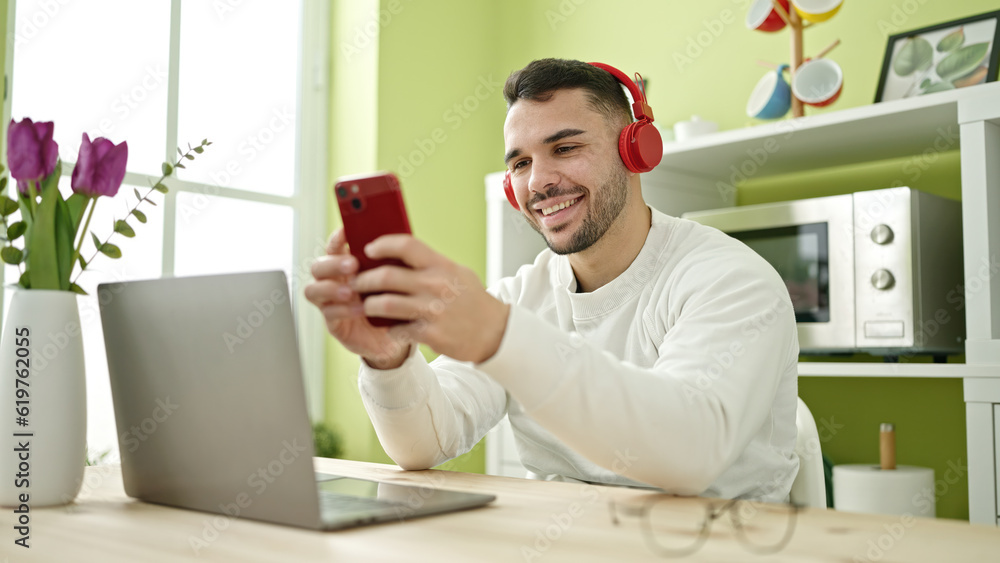 Young hispanic man using smartphone and headphones sitting on table at dinning room