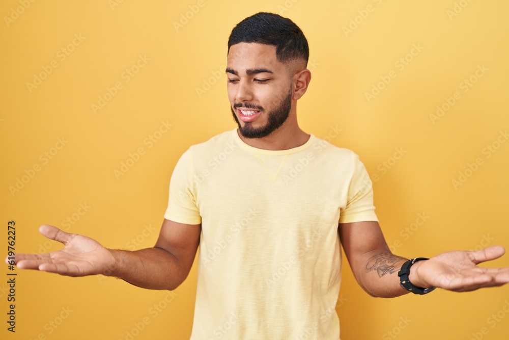 Young hispanic man standing over yellow background smiling showing both hands open palms, presenting and advertising comparison and balance