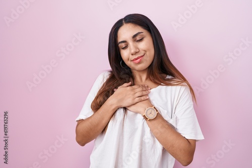 Young arab woman standing over pink background smiling with hands on chest with closed eyes and grateful gesture on face. health concept.