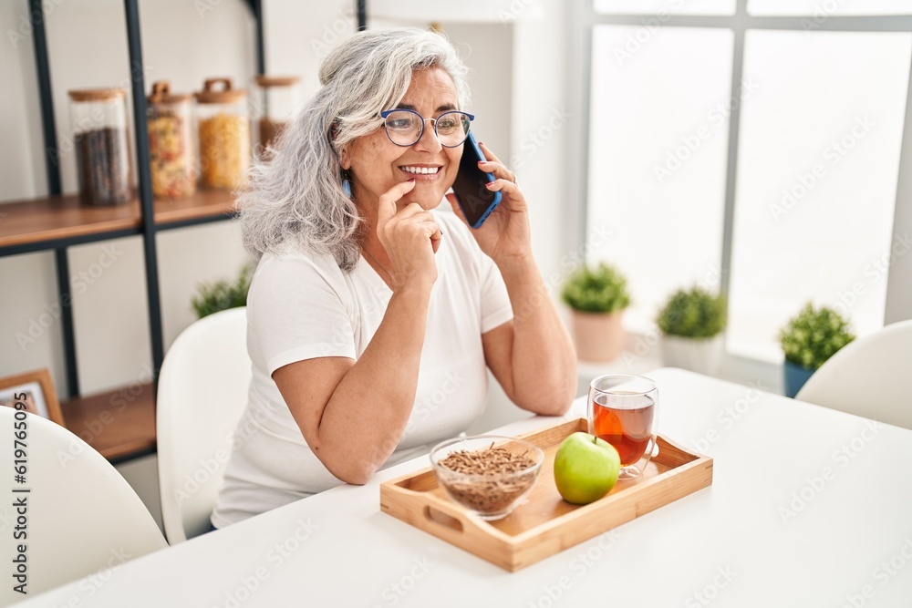 Middle age woman having breakfast talking on the smartphone at home