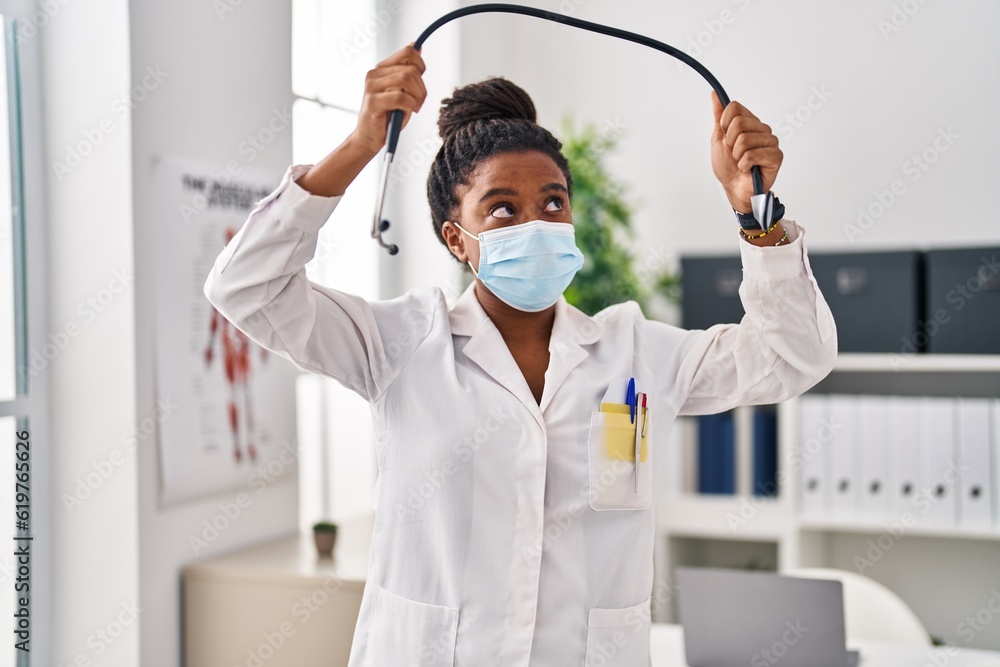 Naklejka premium African american woman wearing doctor uniform and medical mask holding stethoscope at clinic