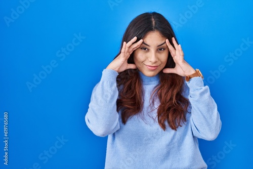 Hispanic young woman standing over blue background trying to open eyes with fingers, sleepy and tired for morning fatigue