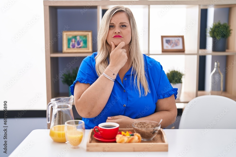 Caucasian plus size woman eating breakfast at home looking confident at the camera with smile with crossed arms and hand raised on chin. thinking positive.