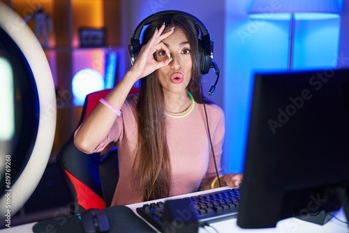 Young hispanic woman playing video games doing ok gesture shocked with surprised face, eye looking through fingers. unbelieving expression.