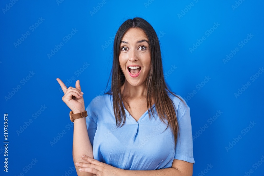 Young brunette woman standing over blue background with a big smile on face, pointing with hand finger to the side looking at the camera. Young brunette woman standing over blue background with a big smile on face, pointing with hand finger to the side looking at the camera.