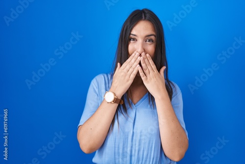 Young brunette woman standing over blue background laughing and embarrassed giggle covering mouth with hands, gossip and scandal concept