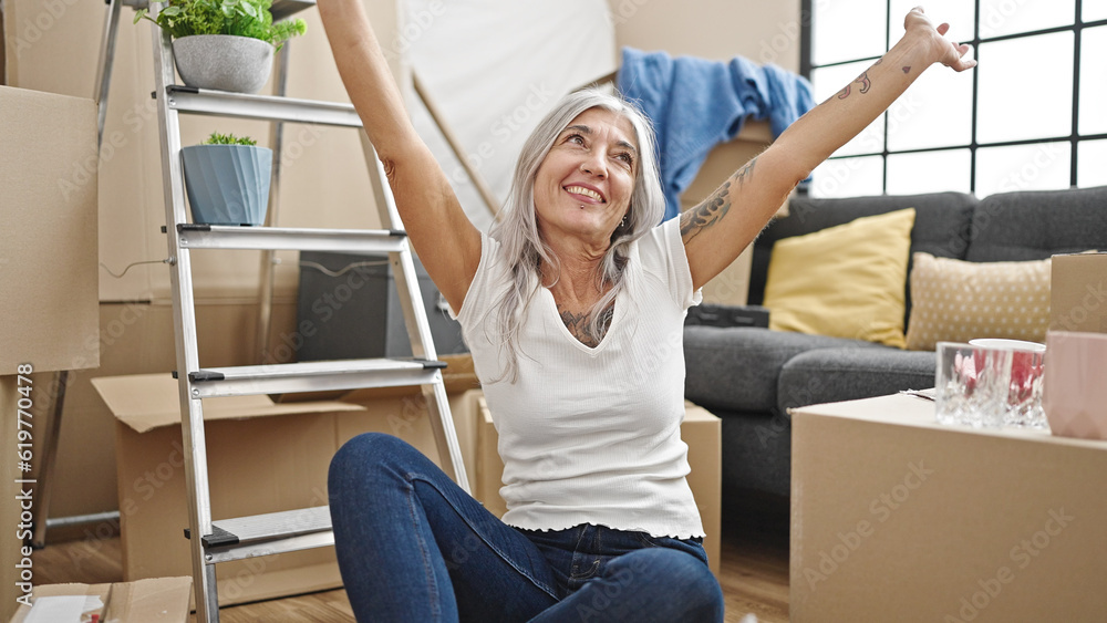 Middle age grey-haired woman sitting on floor with winner expression at new home Middle age grey-haired woman sitting on floor with winner expression at new home