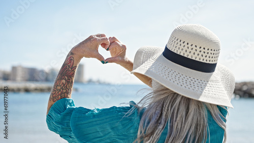 Middle age grey-haired woman doing heart gesture with hands at beach