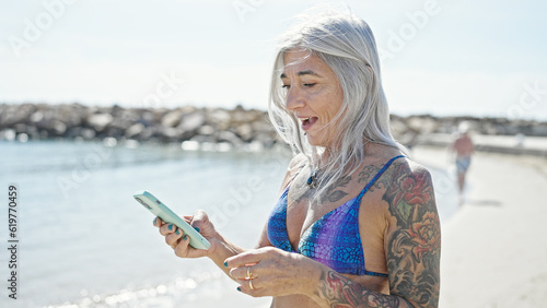Middle age grey-haired woman tourist wearing bikini using smartphone at beach