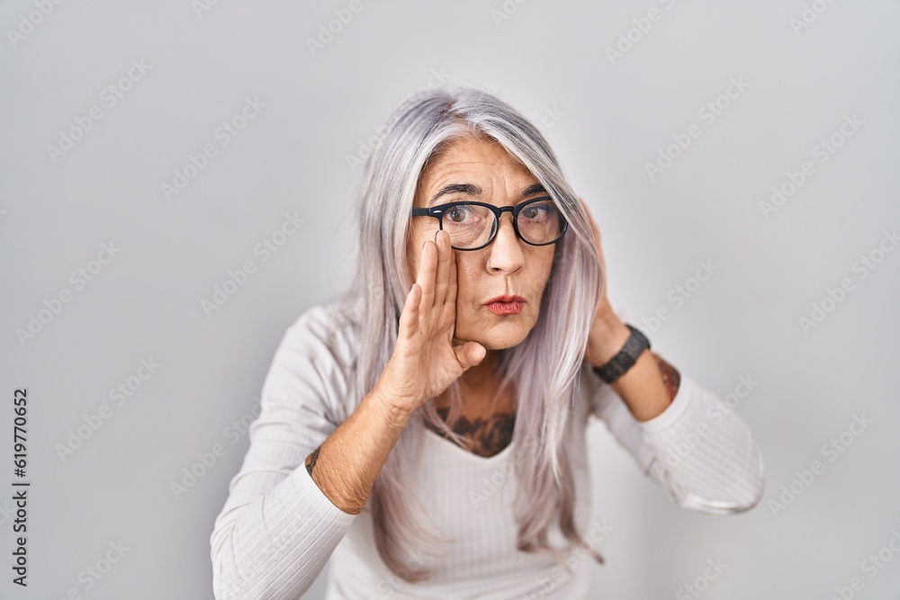 Middle age woman with grey hair standing over white background hand on mouth telling secret rumor, whispering malicious talk conversation Middle age woman with grey hair standing over white background hand on mouth telling secret rumor, whispering malicious talk conversation