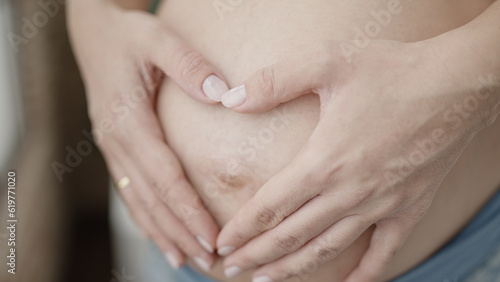 Young pregnant woman doing heart gesture with hands on belly at bedroom