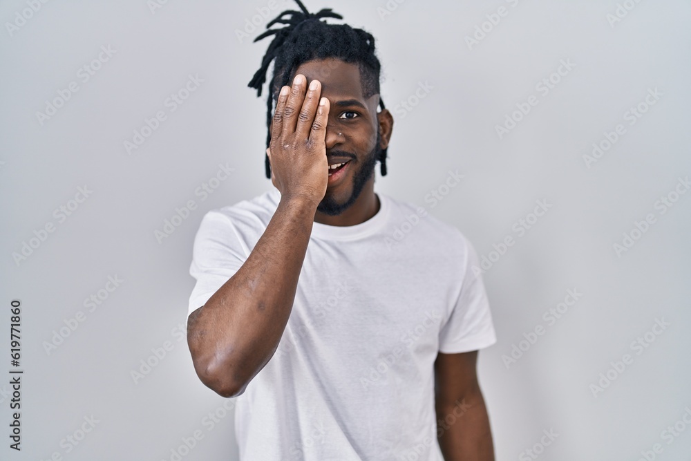 African man with dreadlocks wearing casual t shirt over white background covering one eye with hand, confident smile on face and surprise emotion. African man with dreadlocks wearing casual t shirt over white background covering one eye with hand, confident smile on face and surprise emotion.