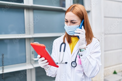 Young caucasian woman doctor wearing medical mask using touchpad talking on smartphone at hospital