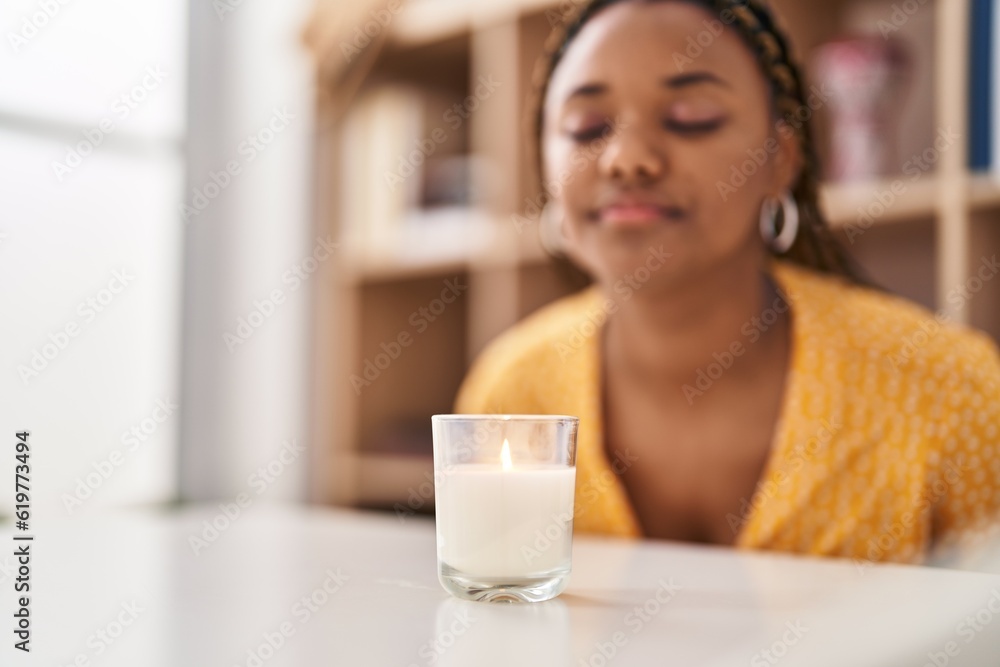 African american woman sitting on table smelling aromatic candle at home African american woman sitting on table smelling aromatic candle at home