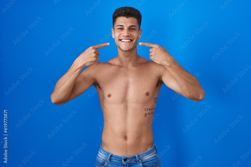 Young hispanic man standing shirtless over blue background smiling cheerful showing and pointing with fingers teeth and mouth. dental health concept.