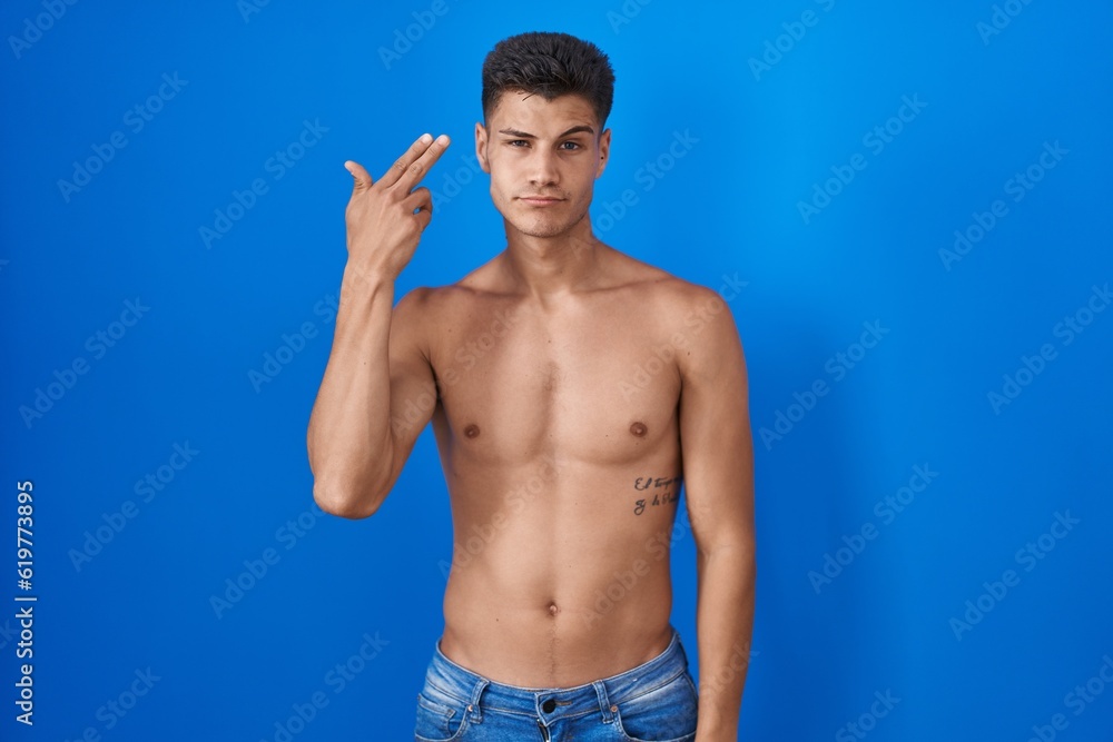 Young hispanic man standing shirtless over blue background shooting and killing oneself pointing hand and fingers to head like gun, suicide gesture.