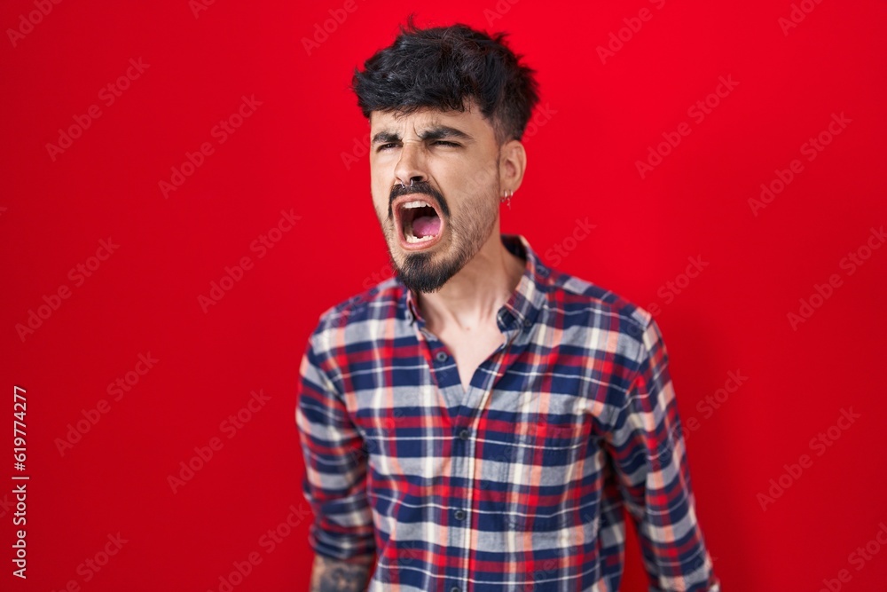 Young hispanic man with beard standing over red background angry and mad screaming frustrated and furious, shouting with anger. rage and aggressive concept. Young hispanic man with beard standing over red background angry and mad screaming frustrated and furious, shouting with anger. rage and aggressive concept.