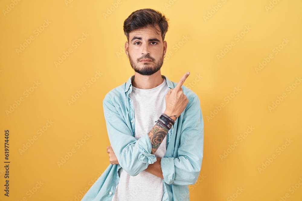 Young hispanic man with tattoos standing over yellow background pointing with hand finger to the side showing advertisement, serious and calm face Young hispanic man with tattoos standing over yellow background pointing with hand finger to the side showing advertisement, serious and calm face